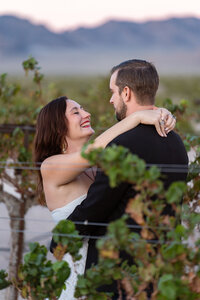 A woman smiles at a man with her arms around his neck in a vineyard