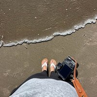 A picture of Danielle Rose Photography taking a photo of her gear from a low angle at Cannon Beach, Oregon, showcasing her travel elopement photographer tools and the beautiful coastal scenery around her.