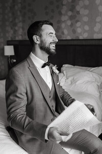 A groom sits on the edge of a hotel bed, laughing with pure joy as he reads a handwritten letter from his bride on the morning of their wedding day. Dressed in a tailored gray suit with a black bow tie and a delicate floral boutonniere pinned to his lapel, he radiates excitement and emotion. The soft, natural light from a nearby window highlights his genuine smile, while the slightly rumpled bed and patterned wallpaper in the background add a sense of warmth and intimacy to the moment. The black-and-white tone of the image enhances the timeless feel, capturing a fleeting yet deeply meaningful pre-ceremony memory.