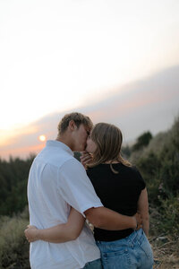 A couple share a kiss outside on a hill in Rexburg, Idaho. In the distance the sun is setting over the trees.