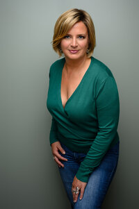 A professional headshot of a confident woman with short blonde hair, wearing a green sweater and jeans against a muted green background. Captured by Leslie Q Photography, specializing in modern headshots and personal branding portraits.