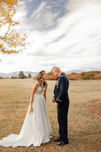 A bride in a white gown and a groom in a military uniform stand close, smiling at each other under a partly cloudy sky. Captured by a Colorado Wedding Photographer, the landscape is an open field with autumn leaves scattered on the ground, painting an enchanting backdrop.