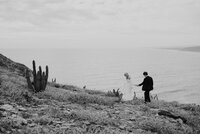 Elegant black and white image of a bride and groom walking hand in hand along a grassy cliffside, overlooking a vast, serene ocean. The bride’s dress flows as the couple takes careful steps together in the coastal breeze.
