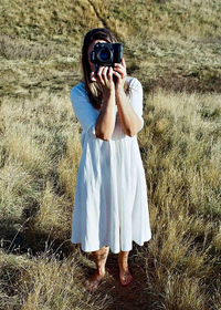 woman wearing a white dress standing in a field holding a camera up to her face