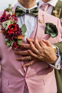 Two grooms are embracing. One is standing in front of the other with their arms together. The groom in front is wearing a light pink suit with a corsage of flowers covering the lapel. 