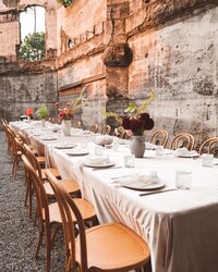 A photo of a wedding reception table set up before guests arrive. It is a very long rectangular table with cream-colored velvet table cloth, light bentwood chairs, white and gold place settings, and bold, colorful flower arrangements spaced out down the middle of the table. There is a beautiful stone wall behind the table. The setting is The Ruins event venue in Hood River, Oregon.