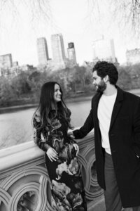 Black-and-white photo of an engaged couple posing together on the iconic Bow Bridge in Central Park, with the New York City skyline in the background, capturing a romantic moment during their engagement session.