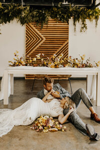 Bride and groom kiss laying on venue ground surrounded by flowers