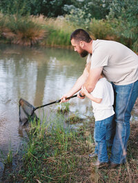 A dad helps his young son fish by a pond, representing a candid moment captured during a family photography session. 