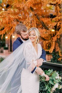 Groom embracing bride from behind with golden leaves in background