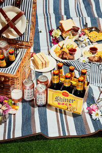 A vibrant and inviting picnic setup featuring a six-pack of Shiner Bock, cans of Underwood wine, and an assortment of picnic foods, all laid out on a striped blanket. Photographed by Mica McCook, this image captures the joy of outdoor gatherings and relaxed dining.