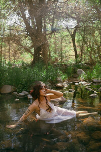 A woman in a white gown sits in a stream surrounded by tall green grass and trees