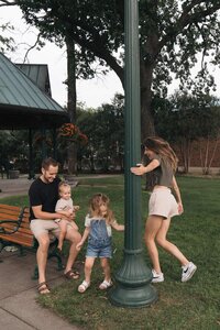 Cinematic wedding photographer twirls around lamppost with oldest daughter while youngest daughter and husband look on from park bench