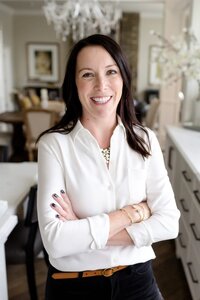 A woman in a crisp white blouse and gold accessories stands confidently with arms crossed in an elegant, well-designed home interior. A polished and professional brand headshot for luxury business owners.