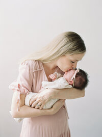 mother wearing pink dress kisses newborn baby girl on forehead captured by Seattle Newborn and Maternity Photographer