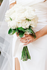 Close-up of a bride holding a bouquet of white roses tied with a satin ribbon, highlighting her oval-cut diamond engagement ring and simple bracelet. Captured in Corinth, Texas.