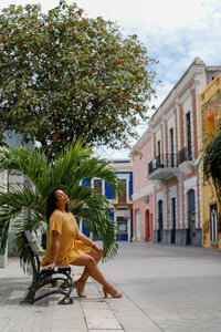 woman sitting on bench outside under a tree and across from colorful building