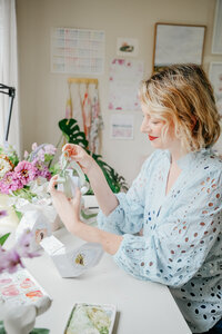 Jess Meyer at a sunlit desk arranging printable treat boxes and painting in her studio.