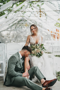 bride sitting in a chair holding a bridal bouquet while groom kisses her leg