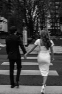 Candid black and white photograph of a couple crossing a street together during an engagement session in downtown Portland, Oregon, captured from behind to highlight their natural connection in an urban Pacific Northwest setting.