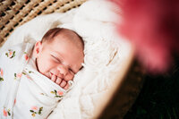 newborn baby swaddled in white blanket with pink flowers on it.  Baby has one hand out of the swaddle and in her smiling mouth.  Photo taken during Philadelphia newborn photography session