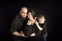 A family of four is posed against a black background. The father stands on the left, with a beard and a black shirt. The mother in the center holds a swaddled baby, wearing a headband. A young boy stands to the right, leaning on the mother and smiling—captured beautifully by Jacksonville newborn photographer.