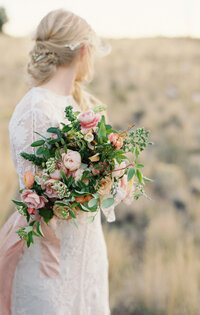 Beautiful bride in an up-tie hairdo looks back into the wide brown pasture with a flower bouquet in her hand.