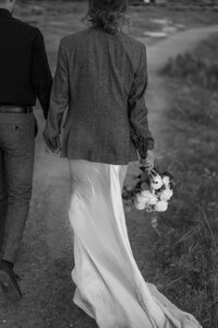 Bride is wearing a silk gown with long flowing train. She holds her long stemmed white roses in one hand and her grooms hand in the other. The bride and groom walk down a path to the Teton Mountain Range in Jackson Hole Wyoming. Photographed by Jackson Hole LUXURY DESTINATION wedding photographer, Magnolia Tree Photo Company