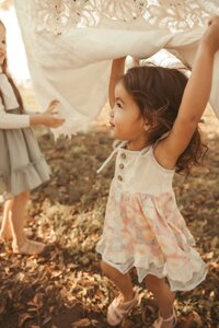 Beautiful little girl twirling with flowing white blanket over her head and her arms raised. She is wearing a flower dress and cute pink sandals.