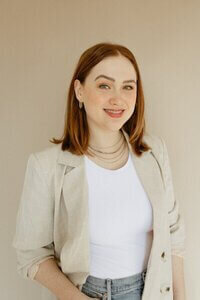 A stylish woman, Caitlin Hart, dressed in a white shirt and denim jacket, showcasing a relaxed and confident demeanor.

