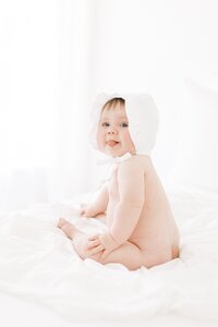 6 month old baby girl with white bonnet sticking tongue out at mom during baby milestone photography session with Amanda Carter