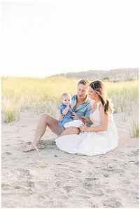 A family of three, mother, father, and baby son sit on the beach during family photos.