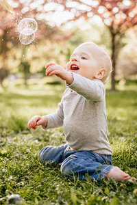A toddler sits on the grass, joyfully reaching for bubbles floating in the air.