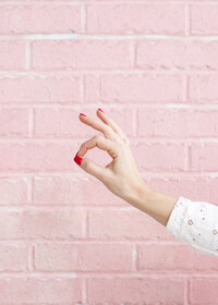 Woman giving a-okay hand sign with red nail polish on pink brick background