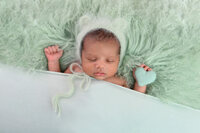 newborn baby laying on his back on a mint green background with a mint green bear hat and holding a mint green heart for his Burlington, Ontario newborn photographer session