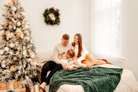 Parents with daughters sit together on bed next to a christmas tree