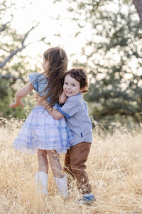 Siblings play hugging each other during a fun golden hour family portrait session at an Oak grove in San Jose