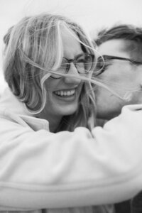 Man kisses his fiance's cheek as the wind whips her hair in front of their faces