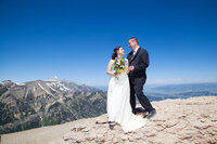 A bride in a lace wedding dress and a groom in a black suit stand atop a mountain in Jackson Hole, gazing at each other with the Teton Range stretching behind them under a bright blue sky.