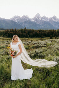photo of bride standing in an open field while wearing her wedding gown and holding her bridal bouquet with mountains in the background taken by Jackson Hole micro wedding photographer