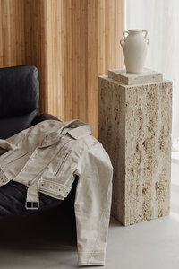 A black leather sofa in a light, minimalist room. A beige jacket is draped over the armrest, and a marble pillar with a white clay vase sits beside it. Light wood cladding covers the wall, with soft natural light filtering through white net curtains.