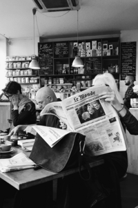 Black and white image of people sitting in a cafe, reading newspapers and enjoying coffee, with shelves of coffee bags and a chalkboard menu in the background.