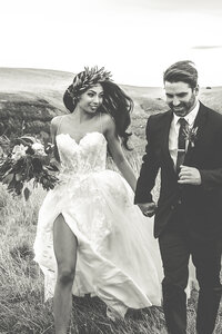 Black and White photograph of a bride and groom running in a field looking happy on wedding day.