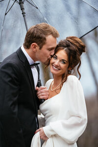 Groom holding a clear umbrella while kissing the cheeks of his bride