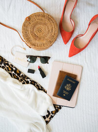 Stylized image of passport and travel accessories sitting on bed with sunglasses and kindle