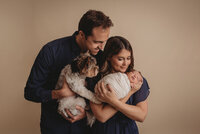 Family of 4 portrait with Atlanta, Georgia newborn photographer family is standing up wearing neutral pinks, browns and creams, with mom holding newborn  baby girl and mom, dad and 2 year old sister are snuggled in close looking at baby and smiling at her.