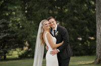 A bride and groom hug and smily for their wedding photos  in front of green trees