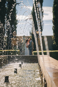 Bride and groom kiss in grand garden as water from a fountain arcs in front of them