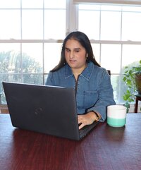Client Ayesha sitting at table on her laptop with mug next to laptop