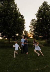 A family portrait of the parents standing together in a picturesque Vermont field while the kids run around them at golden hour.